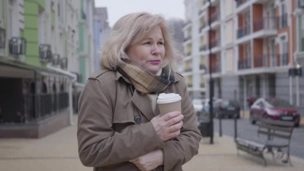 Una mujer caucásica congelada bebiendo café en la calle de la ciudad. Jubilado de mediana edad esperando a alguien en el día de otoño al aire libre. Estilo de vida, ocio, tiempo, estación . — Vídeo de stock