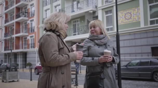 Красивые пожилые кавказские женщины прогуливаются по городской улице, пьют кофе и разговаривают. Две пенсионерки гуляют и болтают на улице. Стиль жизни, досуг, сплетни, отдых . — стоковое видео