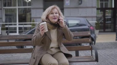 Heyecanlı, son sınıf beyaz kadın portresi telefonda dışarıda bankta oturuyormuş gibi konuşuyor. Neşeli emekli, sonbahar gününü şehir caddesinde geçiriyor. Boş zaman, keyif, yaşam tarzı, iletişim.