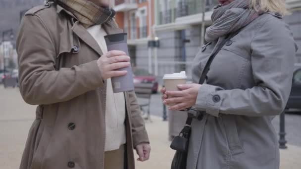 Duas mulheres caucasianas de meia-idade irreconhecíveis a conversar ao ar livre. Reformados elegantes em casacos de outono em pé na rua da cidade com xícaras de café, conversando e gesticulando. Lazer, descanso, estilo de vida . — Vídeo de Stock