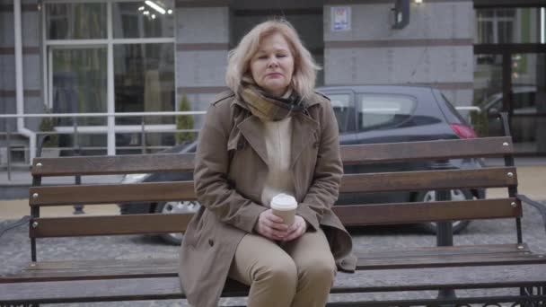 Retrato de bela mulher caucasiana de meia-idade bebendo café e pensando na rua da cidade. Um aposentado atencioso, sentado sozinho no banco ao ar livre. Solidão, estilo de vida, pensão, tristeza . — Vídeo de Stock