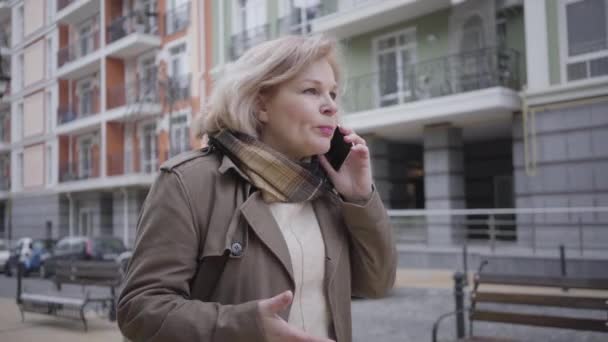 Şehir caddesinde yürüyen ve telefonda konuşan olgun, zarif beyaz kadın portresi. Yaşlı bayan mikrofona bağırıp el kol hareketi yapıyor. Yaşam tarzı, iş hayatı, stres. — Stok video