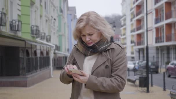 Porträtt av stressad äldre kaukasisk kvinna står på utländsk stadsgata med smartphone försöker hitta vägen. Förlorade pensionerad turist tittar runt och söker riktning i mobilappen. Turism. — Stockvideo