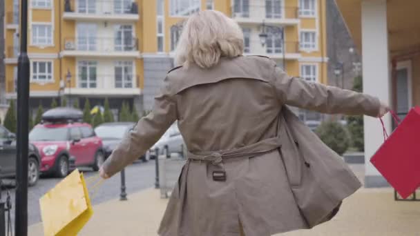 Portret van vrolijke blanke vrouw van middelbare leeftijd in herfstkleding en zonnebril draaiend en dansend met boodschappentassen buiten. Vreugdevolle gepensioneerde genieten van vrije tijd op straat. Levensstijl. — Stockvideo