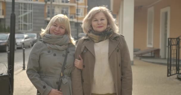 Két idősebb kaukázusi nő portréja, akik a szabadban pózolnak. Boldog nyugdíjasok mosolyognak a kamera előtt a város utcáján. Szabadidő, nyugdíj, nyugdíj, életmód. Mozi 4k Prores Hq. — Stock videók
