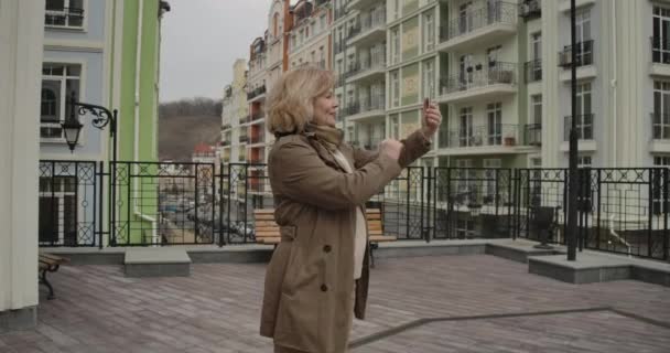 Θετική ηλικιωμένη λευκή γυναίκα να χαιρετάει και να μιλάει στην κάμερα selfie έξω. Χαρούμενη συνταξιούχος απολαμβάνοντας την ημέρα του φθινοπώρου στην ξένη πόλη. Τουρισμός, ταξίδια, τρόπος ζωής. Κινηματογράφος 4k Prores Hq. — Αρχείο Βίντεο