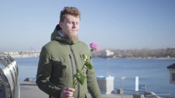 Портрет молодого кавказького рудого чоловіка з вусами і бородою, який чекає на дорослу троянду. Хлопець стоїть на березі річки в сонячний осінній день і пахне квіткою. Романс, любов. — стокове відео