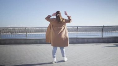 Neşeli genç tombul kadın güneşli bir sonbahar gününde nehir kıyısına doğru yürüyor. Kamera, açık havada eğlenen mutlu beyaz esmer kızı takip ediyor. Yaşam tarzı, mutluluk, yalnızlık.