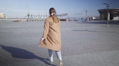 Şehir meydanında gezen ve gülümseyen neşeli beyaz kadın portresi. Kameralar pozitif kızın yürüyüşünü, dönüşünü, güneşli sonbahar gününün tadını çıkarışını gösteriyor. Boş zaman, yaşam tarzı, neşe.