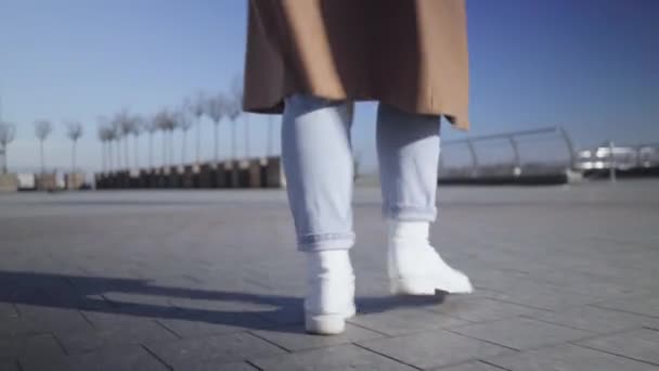 Caméra suivant les jambes féminines en jeans et baskets blanches marchant sur la place de la ville. Jeune femme flânant à l'extérieur par une journée ensoleillée. Mode de vie, vie urbaine, repos, loisirs . — Video