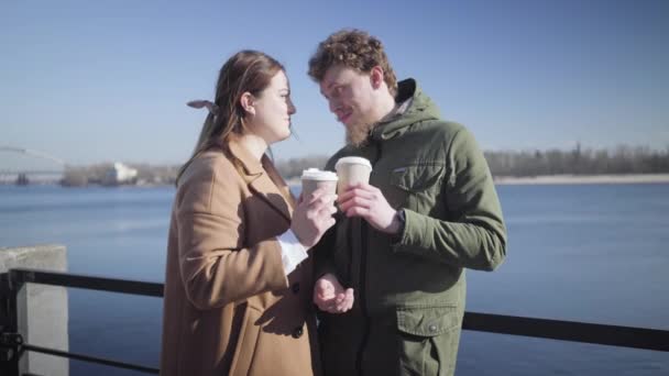 Retrato de una joven pareja enamorada de tomar café en la orilla del río. Feliz hombre y mujer caucásicos citas al aire libre en el día soleado. Chica poniendo la cabeza en los hombres hombro y sonriendo. Felicidad, alegría, amor . — Vídeos de Stock