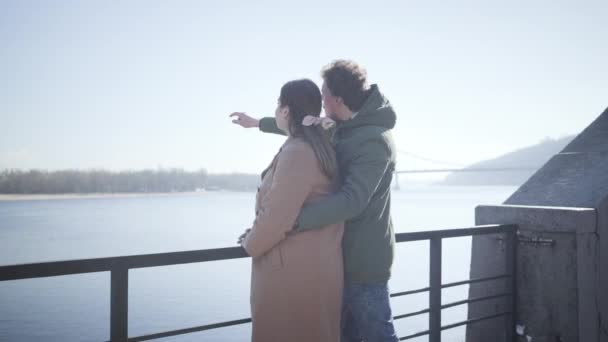 Vista lateral de pareja feliz en el amor charlando en la orilla del río y apuntando a distancia. Joven caucásico hombre y mujer citas al aire libre. Alegría, ocio, romance, estilo de vida . — Vídeos de Stock