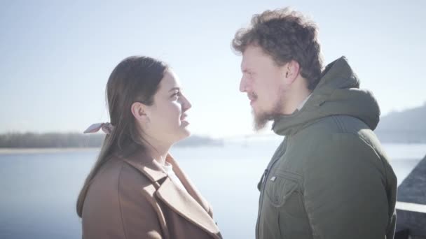 Zijaanzicht van gelukkige blanke man en vrouw die elkaar aankijken met liefde en wrijvende neuzen. Jong stel op de oever van de rivier in het zonlicht en glimlachend. Liefde, romantiek, afspraakjes, eenheid. — Stockvideo