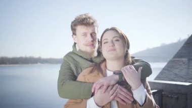 Kameraya bakıp dışarıda gülümseyen mutlu çiftin portresi. Güneşli bir sonbahar gününde nehir kıyısında poz veren beyaz erkekleri ve kadınları sevmek. Boş zaman, aşk, aşk, yaşam tarzı..