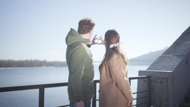 Счастливый молодой кавказский мужчина и женщина придают форму сердцу руками на фоне голубого осеннего неба. Улыбающаяся пара смотрит друг на друга с любовью, как стоя на берегу реки на открытом воздухе. Любовь, романтика — стоковое видео
