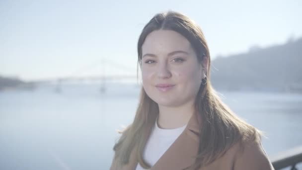 Gros plan portrait de la jolie brune caucasienne en plein soleil. Fille joufflu avec les yeux bruns regardant la caméra et souriant comme debout sur le pont à l'arrière-plan de la rivière calme. Mode de vie . — Video