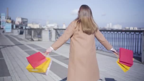 Retrato de mujer gordita joven feliz girando con bolsas de compras en la orilla del río en el día soleado de otoño. Mujer blanca disfrutando de tiempo libre al aire libre. Ocio, estilo de vida, consumismo, compras . — Vídeos de Stock