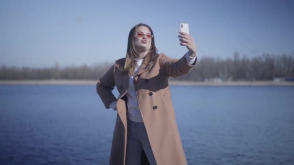 Портрет позитивної молодої жінки, яка надсилає повітряний поцілунок і скаржиться на саморобну камеру на вулиці. Щаслива кавказька дівчина фотографується на березі річки в сонячний день. Радість, дозвілля, спосіб життя, щастя. — стокове відео