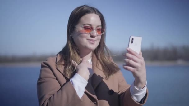 Detailní záběr radostné mladé ženy ve slunečních brýlích mluvící na selfie kameru na smartphonu venku. Pozitivní usmívající se běloška, která má videohovor, jak odpočívá na břehu řeky za slunečného podzimního dne. Životní styl. — Stock video