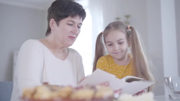 Vuxen brunett vit kvinna och liten flicka diskuterar recept på pannkakor för Shrove tisdag. Positiv mormor och barnbarn matlagning söt dessert för semester. Shrovetide, livsstil. — Stockvideo
