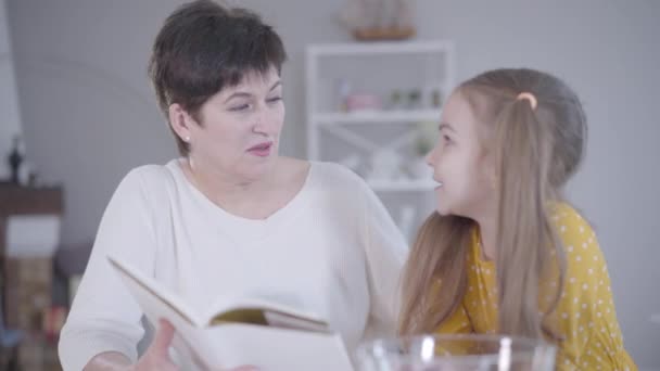 Mujer adulta segura hablando con su nieta en la cocina y leyendo la receta. Linda niña caucásica ayudando a la abuela a cocinar panqueques para Shrovetide. Estilo de vida, Hornear, Martes de Carnaval . — Vídeo de stock