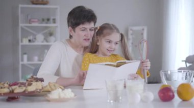 Güzel küçük bir kızın portresi. Diz çöküp yemek tarifini dinliyor. Beyaz bir büyükanne torunuyla Shrove salısı için krep pişiriyor. Shrovetide, yaşam tarzı, pişirme