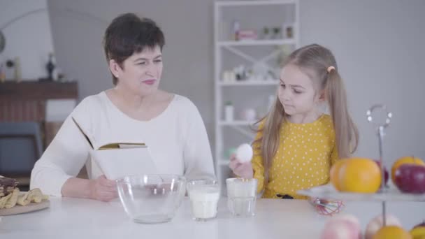 Portret van een ijverig meisje dat oma helpt met het bakken van pannenkoeken voor Shrove Tuesday. Mooi blank kind dat eieren, bloem en melk verzamelt als vrouw die ingrediënten uit recept leest. — Stockvideo