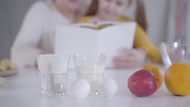 Närbild av mjölk, ägg, mjöl och frukt på bordet med suddiga människor läser recept på bakgrunden. Kaukasiska liten flicka matlagning pannkakor för Shrovetide med mormor. Shrove tisdag. — Stockvideo