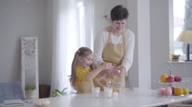 Neşeli beyaz kız içeride yemek pişirmede büyükanneme yardım ediyor. Gülümseyen küçük torun kaseye yumurta kırıyor. Shrove Salısı için aile krepleri pişiriyoruz. Yaşam tarzı, mutfak, Shrovetide.