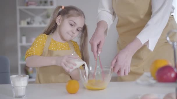Cute Caucasian brunette girl pouring milk into bowl with eggs. Unrecognizable adult woman beating ingredients for baking of pancakes. Granddaughter helping grandmother with cooking. — Stock Video