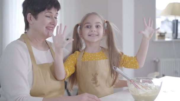 Menina bonito sorrindo para a câmera na cozinha como avó rindo. Criança caucasiana em avental sujo com ingredientes que ajudam a mulher positiva a assar panquecas na terça-feira Shrove. Comida, Shrovetide . — Vídeo de Stock