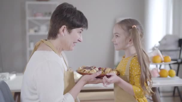 Πλευρική άποψη του ευτυχισμένη γιαγιά και εγγονή κρατώντας πιάτο με ψημένα νόστιμα τηγανίτες και τρίψιμο μύτες. Χαρούμενη γυναίκα και κορίτσι ψήνουν επιδόρπιο για το Shrove Τρίτη. Τρόπος ζωής, χαρά, Shrovetide. — Αρχείο Βίντεο