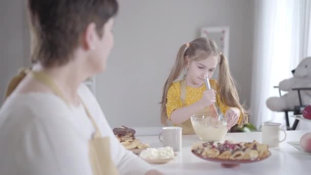 Allvarlig liten flicka slår ingredienser i skålen och ler till suddig kvinna i förgrunden. Barnbarn hjälper mormor att baka pannkakor till Shrove Tuesday. Livsstil, lycka, Shrovetide. — Stockvideo