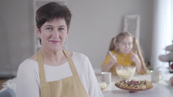 Ritratto ravvicinato di felice donna adulta caucasica che sorride alla telecamera mentre la bambina sfocata lecca le dita con ingredienti dolci per la cottura. Positivo nonna e nipote cucina . — Video Stock