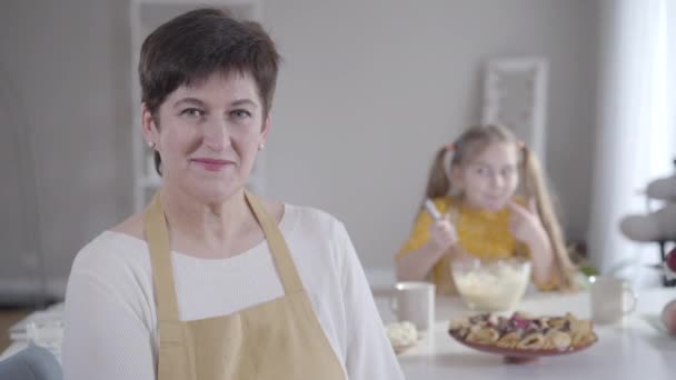 Close-up portrait of happy Caucasian adult woman smiling at camera as blurred little girl licking fingers with sweet ingredients for baking. Positive grandmother and granddaughter cooking. — 비디오