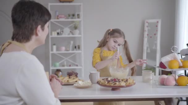 Schattig blank meisje met duimen omhoog en glimlachend naar onherkenbare oma. Mooie kind slaan ingrediënten voor het bakken in kom binnen. Shrovetide, koken, levensstijl, vreugde. — Stockvideo