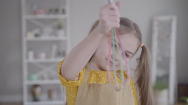 Porträt eines charmanten kleinen Mädchens mit braunen Augen, das Schneebesen hält. Der Fokus wechselt vom Kindergesicht zum bunten Kochgeschirr. Lebensstil, Kochen, Backen, Fastnacht. — Stockvideo