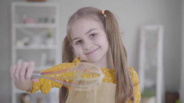 Menina caucasiana pouco agradável com olhos castanhos sorrindo para a câmera e levantando batedor de mistura para assar. Bonita criança a cozinhar dentro de casa. Shrove terça-feira, assar, panquecas, Shrovetide . — Vídeo de Stock