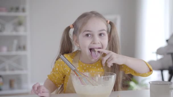 Retrato de bonito alegre caucasiano menina degustação batido ingredientes da tigela. Criança morena alegre com olhos castanhos mergulhando os dedos em mistura doce e lambendo-os. Cozinhar, padaria, Shrovetide . — Vídeo de Stock