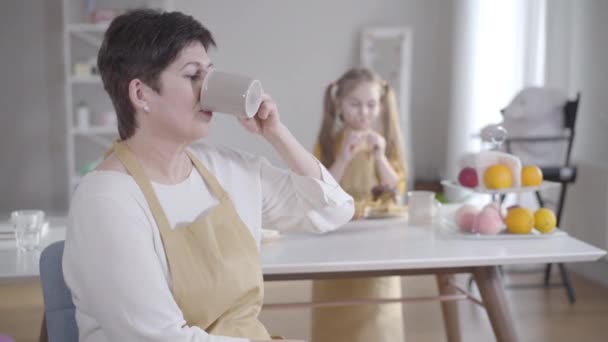 Positiv brunett Kaukasiska kvinna dricker te och ler mot kameran som suddig liten flicka äter pannkakor i bakgrunden. Glad mormor tillbringar Shrove tisdag med barnbarn. — Stockvideo