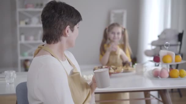 Brunette adult woman looking back at granddaughter eating delicious pancakes, turning to camera and smiling. Happy grandmother resting with little cute girl on Shrove Tuesday. Lifestyle, unity. — Stock Video