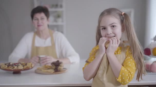 Porträtt av söt liten flicka som äter pannkaka med suddig kaukasisk mormor ler mot bakgrunden. Brunett söt barn med bruna ögon smaka läcker dessert. Matlagning, Shrove tisdag. — Stockvideo