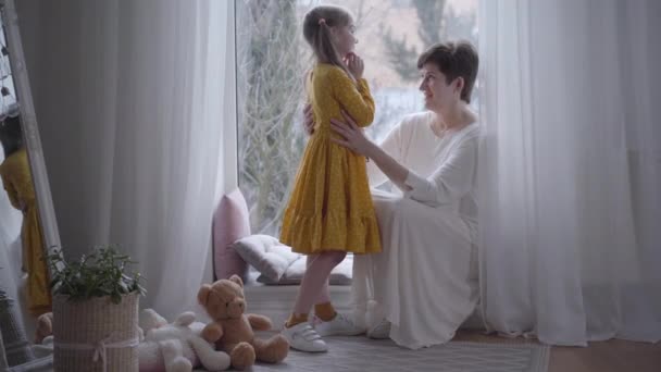 Elegante mujer caucásica en vestido blanco abrazando a la linda niña en traje de puntos amarillos. Hermosa abuela y nieta pasar el día juntos en el interior. Estilo de vida, alegría, unidad . — Vídeos de Stock