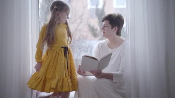 Chica bastante caucásica que viene a la abuela sentada en el alféizar de la ventana con el libro. Nieta sonriente y abuela leyendo en interiores. Aficion, estilo de vida, inteligencia, ocio . — Vídeo de stock