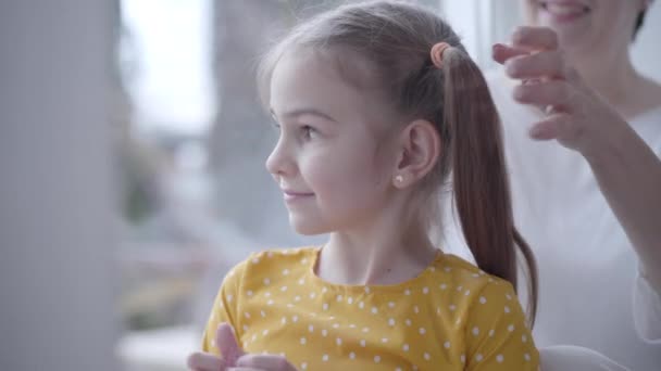 Nahaufnahme Porträt eines charmanten kleinen Mädchens, das mit Spielzeugschmuck spielt, als unkenntliche Frau, die ihre Pferdeschwänze im Hintergrund bindet. Glückliche Enkelin genießt Zeit mit Oma im Haus. — Stockvideo