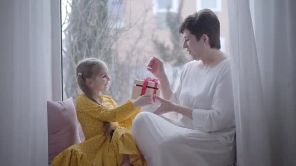 Glada kaukasiska flicka ger gåva till positiv mormor. Vuxen brunett kvinna och söt liten barn chattar inomhus sitter på fönsterbrädan. Fritid, glädje, livsstil, lycka. — Stockvideo