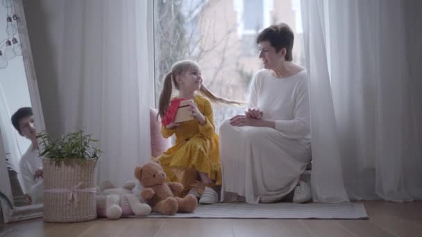 Wide shot, elegant adult woman touching granddaughters shoulder, child turning to her with gift in hands. Portrait of cute girl giving present to grandmother indoors. Joy, leisure, lifestyle, unity. — 비디오