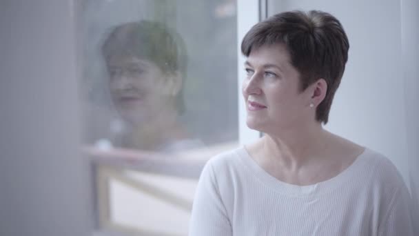 Portrait of thoughtful Caucasian adult woman looking out the window. Beautiful senior lady smiling and thinking at home. Lifestyle, retirement, leisure, resting. — Stock Video