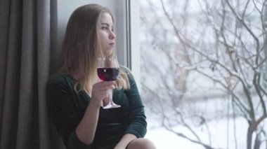 Düşünceli genç beyaz kadın pencereden dışarı bakıyor ve camdan kırmızı şarap içiyor. Yalnız, güzel bir kız, evde tek başına bir gün geçiriyor. Zarafet, yaşam tarzı, yalnızlık, depresyon.