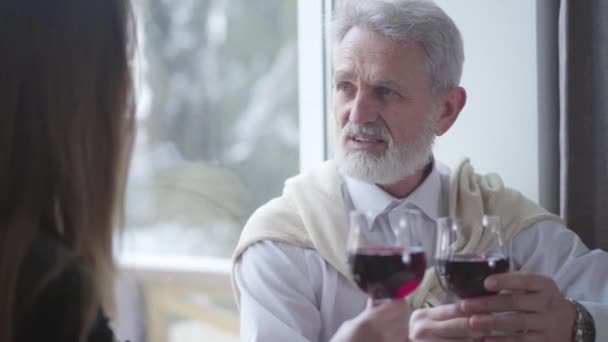 Primer plano retrato de un hombre mayor guapo y seguro hablando con una morena irreconocible y sonriente. jubilado caucásico alegre pasar tiempo con la joven esposa en el interior beber vino tinto . — Vídeo de stock